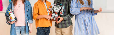 Panoramic shot of schoolkids holding robots and laptop in stem school