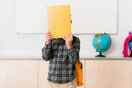Schoolboy covering face with book in classroom of stem school