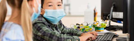 Website header of asian schoolboy in medical mask using computer near classmate in classroomの写真素材