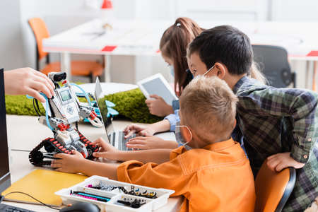 Selective focus of teacher holding robot near multiethnic pupils in medical masks and gadgets in...