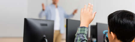 Horizontal crop of asian schoolboy with raised hand sitting near computers in stem school