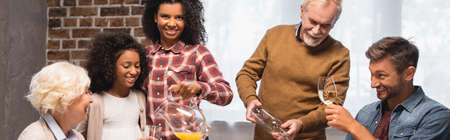 panoramic shot of senior man and african american woman pouring orange juice and white wine during...