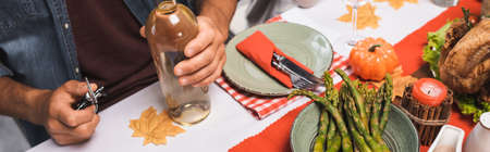 cropped view of man holding bottle of white wine and corkscrew at table served with thanksgiving dinner, horizontal imageの写真素材