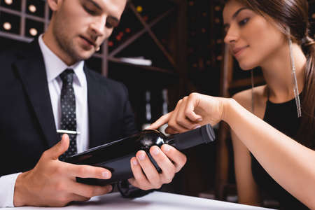 Selective focus of woman pointing at bottle of wine near boyfriend in suit in restaurantの写真素材