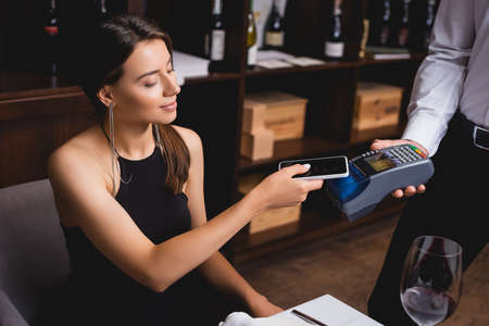 Selective focus of young elegant woman paying with cellphone near waiter with payment terminal in...