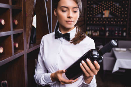 Selective focus of young sommelier in formal wear looking at bottle of wine in restaurantの写真素材
