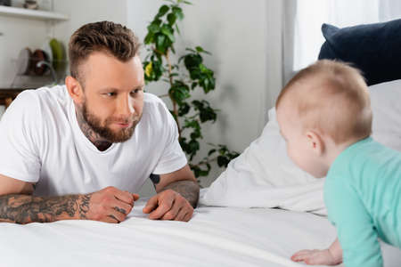 selective focus of tattooed man looking at infant child crawling on bedの写真素材