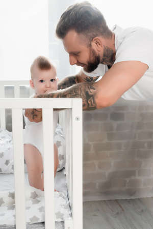 tattooed man supporting infant child standing in cot and holding hand in mouthの写真素材