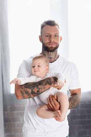 young tattooed man looking at camera while holding joyful infant son in baby romperの写真素材