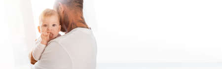 website header of infant boy holding hand in mouth while looking at camera in fathers handsの写真素材