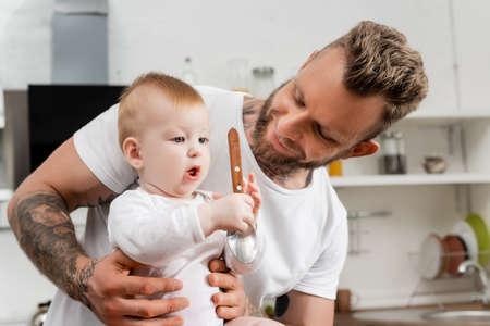 infant boy holding spoon near young, bearded father in kitchen