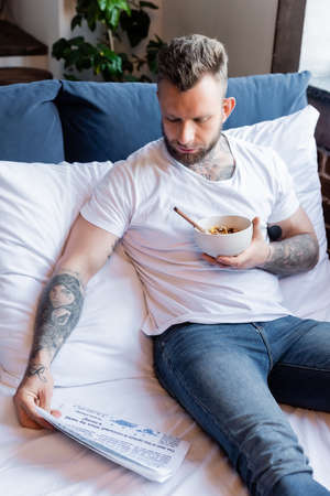 tattooed man in white t-shirt in jeans reading newspaper while having breakfast in bedの写真素材