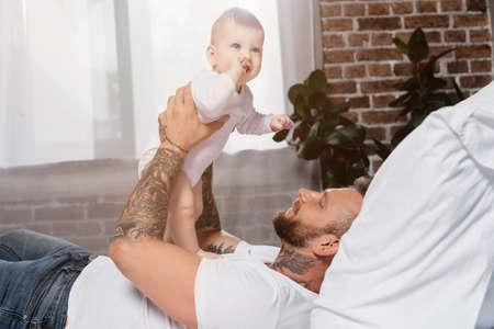 selective focus of young tattooed man holding excited baby boy while lying in bedの写真素材