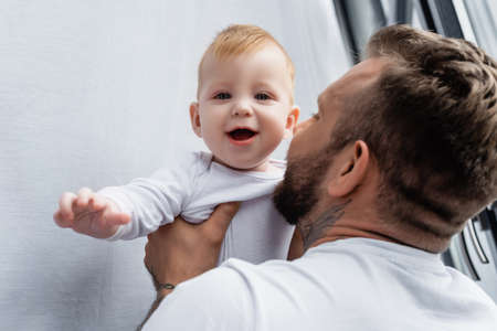 selective focus of young father holding excited infant son looking at camera with open mouth