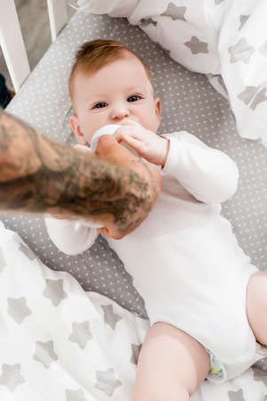 selective focus of tattooed father feeding baby boy lying in cot in baby romper