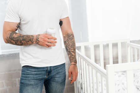 partial view of tattooed man in white t-shirt and jeans holding baby bottle while standing near crib
