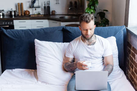 young, tattooed freelancer using laptop in bed while holding cup of coffeeの写真素材