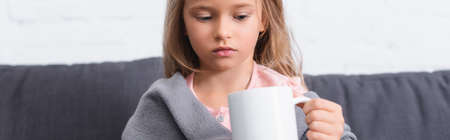 Panoramic shot of ill child in blanket looking at mug at homeの写真素材