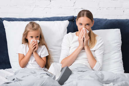 Selective focus of sick mother and daughter holding napkins while having snuffle on bedの写真素材