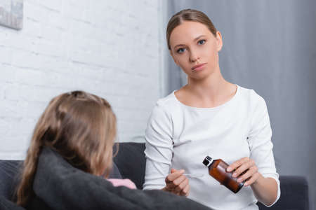 Selective focus of woman looking at camera while holding syrup and spoon near ill daughter on couchの写真素材