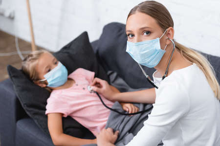 Selective focus of woman in medical mask and stethoscope sitting near child at homeの写真素材