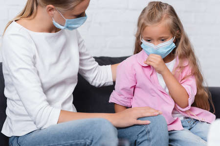 Selective focus of girl in medical mask sitting near mother on couchの写真素材
