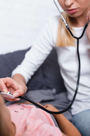 Selective focus of woman using stethoscope near daughter lying on sofa