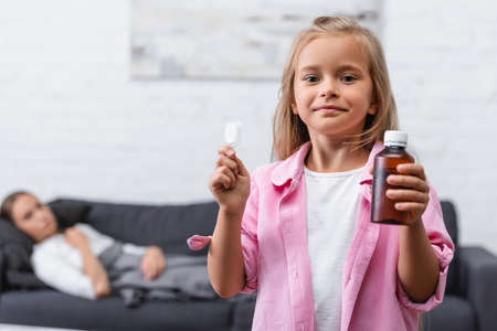 Selective focus of girl holding spoon and bottle of syrup near ill mother in living roomの写真素材