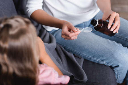 Selective focus of woman pouring syrup in spoon near diseased child on sofa in living roomの写真素材