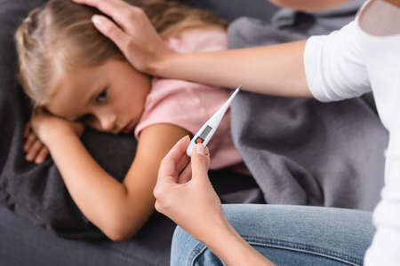 Selective focus of woman holding thermometer and stroking sick daughter on couch