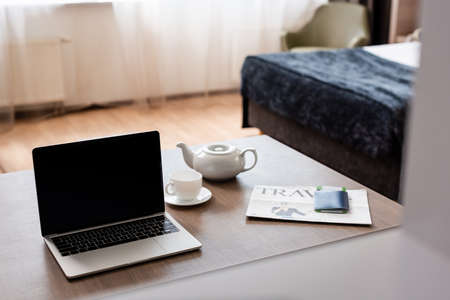 laptop with blank screen near teapot, cup, travel newspaper and passport with air ticket on tableの写真素材