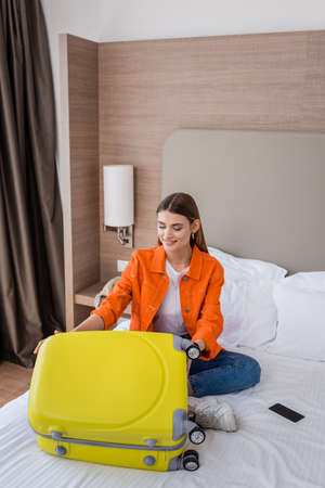 young woman sitting on bed near yellow baggage and smartphone with blank screen in hotelの写真素材
