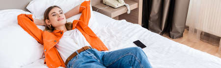 panoramic shot of young woman resting near smartphone with blank screen on bed in hotelの写真素材