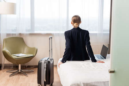 back view of businesswoman sitting on bed near laptop with blank screen and suitcase in hotel roomの写真素材