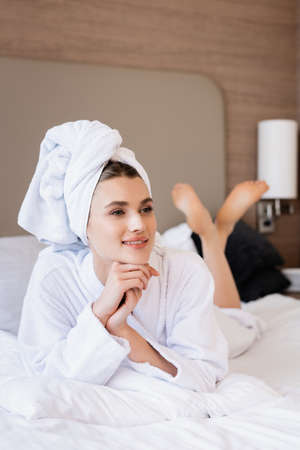 barefoot woman in towel and white bathrobe lying on bed and looking away in hotel roomの写真素材