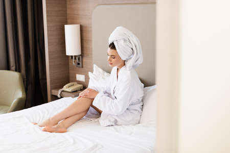 selective focus of barefoot woman in towel and white bathrobe sitting on bed and touching legs in hotel roomの写真素材