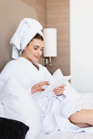 young woman in towel and white bathrobe looking at digital tablet in hotel roomの写真素材