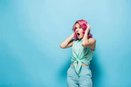 shocked young woman with pink hair and open mouth holding head on blue background