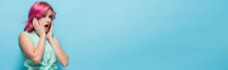 surprised young woman with pink hair and open mouth touching face on blue background, panoramic shot