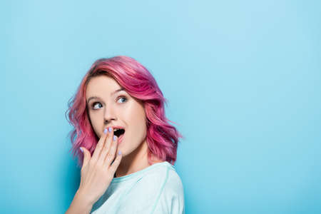 shocked young woman with pink hair covering mouth on blue background