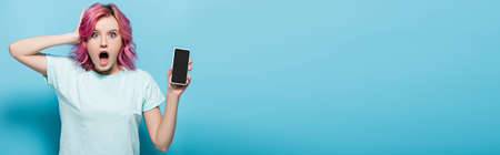 shocked young woman with pink hair holding smartphone with blank screen on blue background,...