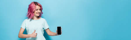 young woman with pink hair holding smartphone with blank screen and showing thumb up on blue background, panoramic shotの写真素材