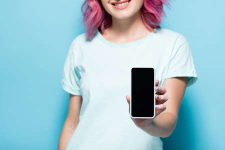 cropped view of young woman with pink hair holding smartphone with blank screen on blue background