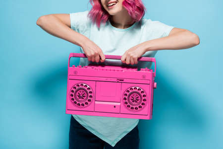 cropped view of young woman with pink hair holding vintage tape recorder on blue background