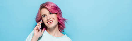 young woman with pink hair talking on smartphone and smiling on blue background, panoramic shot