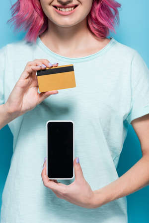 partial view of young woman with pink hair holding credit card and smartphone on blue background