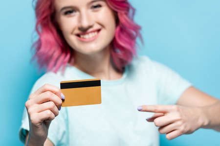 selective focus of young woman with pink hair pointing at credit card on blue background