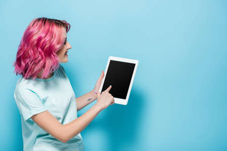 side view of young woman with pink hair using digital tablet on blue background