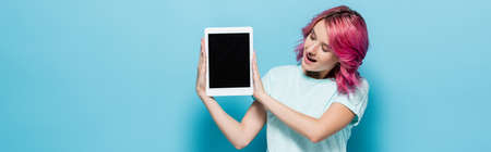surprised young woman with pink hair presenting digital tablet with blank screen on blue background,...