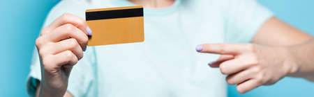 selective focus of young woman pointing at credit card on blue background, panoramic shot
